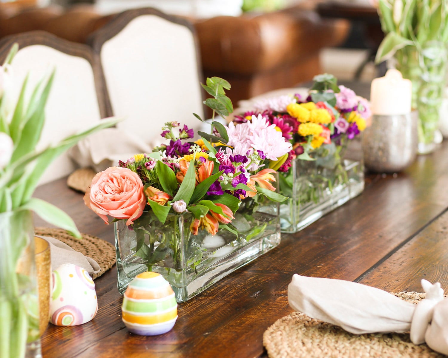 A wooden dining table is styled with colorful spring flower arrangements in rectangular glass vases, surrounded by Easter eggs, candles, and placemats