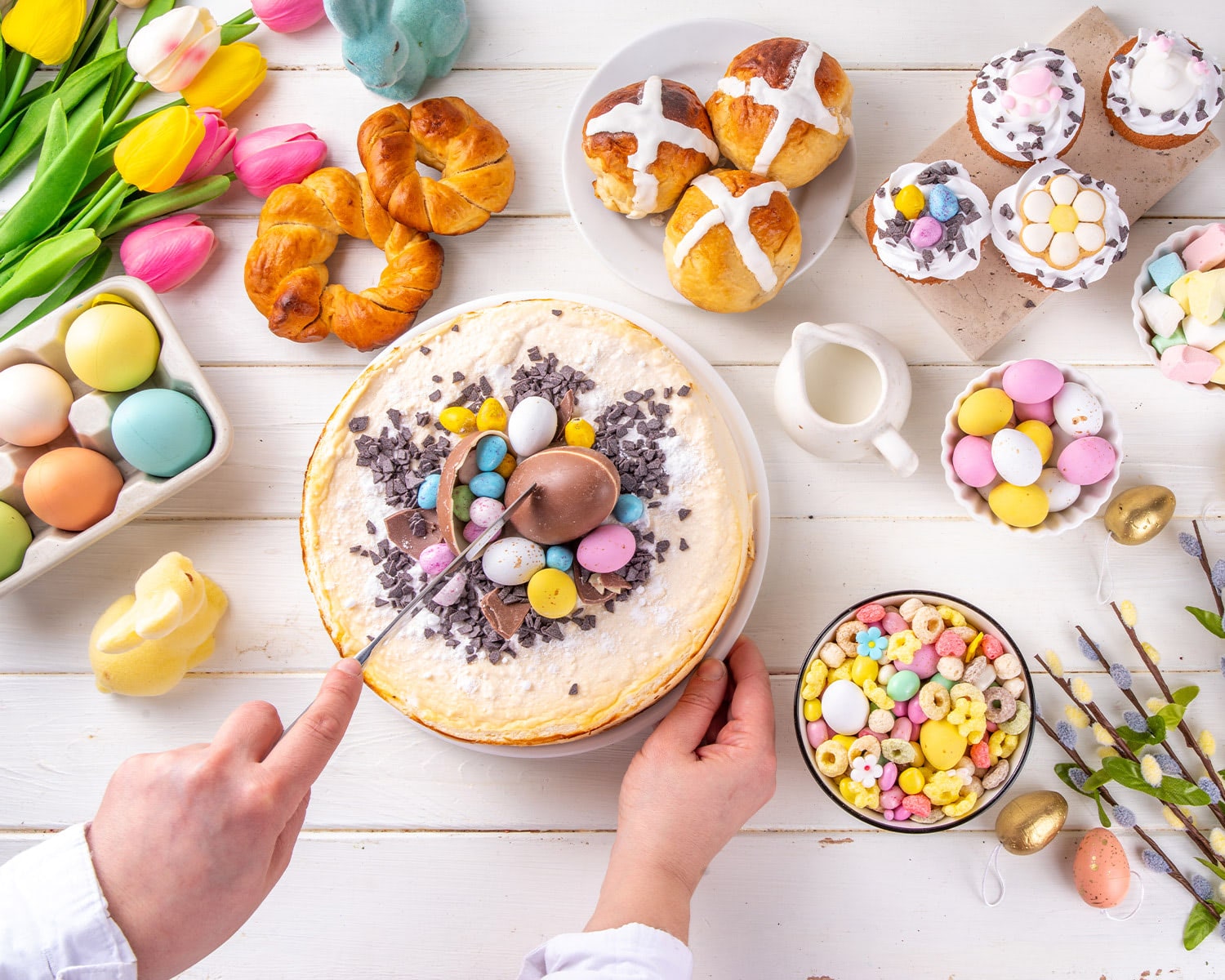 A person slices into Easter cheesecake decorated with chocolate eggs and sprinkles, surrounded by spring pastries, candies, tulips, and dyed eggs